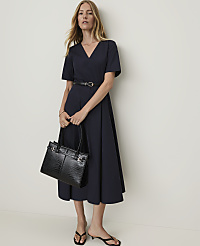 A woman in a navy dress and black sandals holds a black crocodile-embossed handbag in front of a plain white wall. carousel Product Image 1