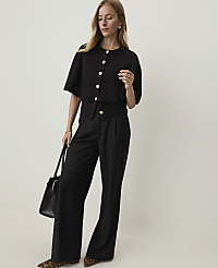 A woman in a black cardigan and wide-leg pants stands against a plain white background, holding a black handbag. carousel Product Image 3