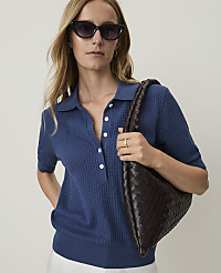 A woman in a blue polo shirt and sunglasses holds a dark brown woven bag in front of a plain white background. carousel Product Image 1