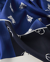 A close-up of a blue silk scarf with a pattern of white horse heads and equestrian tack in the foreground, set against a background of darker blue and white sections of the fabric. carousel Product Image 2