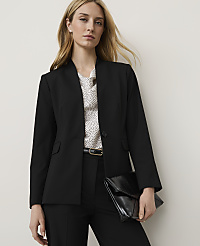 A woman in a black blazer and polka dot blouse holds a black clutch in front of a plain background. carousel Product Image 1