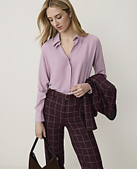 A woman in a lilac blouse and plaid pants stands against a plain white background, holding a dark handbag. carousel Product Image 1