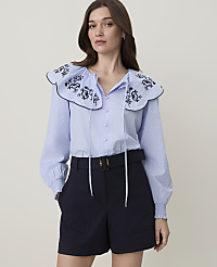 A woman in a blue striped blouse with a large embroidered collar and dark shorts stands against a plain, light background. carousel Product Image 1