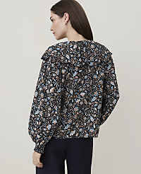 A woman in a dark floral blouse with a ruffled collar stands against a plain, light-colored background. carousel Product Image 2