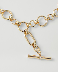 A gold toggle clasp necklace with circular links in the foreground rests on a plain white background. carousel Product Image 2