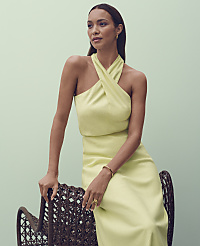 A woman in a light yellow halter dress sits on a wicker chair against a soft green background. carousel Product Image 3