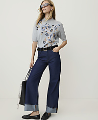 A woman in a grey floral sweater and dark wide-leg jeans stands against a plain white background, holding a black tote bag. carousel Product Image 3