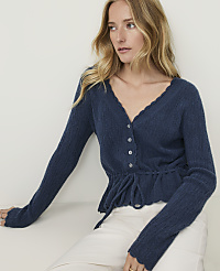 A woman in a blue ruffled cardigan and white pants poses against a plain white background. carousel Product Image 1
