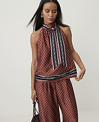A woman in a polka-dot halter top and matching pants stands against a plain, light background, holding a dark handbag.
