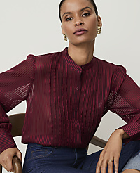 A woman in a burgundy sheer blouse with vertical pleats and dark jeans sits in front of a blurred background. carousel Product Image 1
