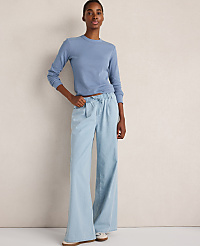 A woman in a blue long-sleeved shirt and wide-leg light blue jeans stands against a plain, textured beige background. carousel Product Image 4