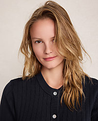 A young woman with blonde hair and a dark blue cardigan smiles gently against a plain, light-colored background. carousel Product Image 2