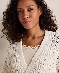 A woman with curly dark hair wears a cream-colored, open-knit cardigan over a cream-colored top with a scalloped neckline, against a soft, blurred background. carousel Product Image 2