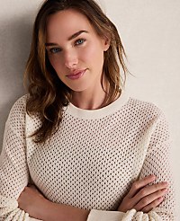 A woman in a cream-colored, textured sweater poses with her arms crossed against a neutral, textured background. carousel Product Image 2