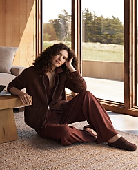 A woman in a brown loungewear set sits on a jute rug in a sunlit room with a wooden table and sofa, while a large window reveals a blurred outdoor landscape. carousel Product Image 2