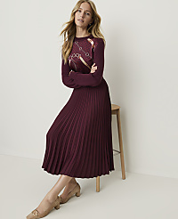 A woman in a burgundy pleated dress with a patterned top sits on a stool against a plain white background. carousel Product Image 3