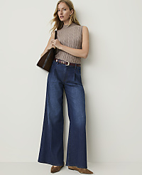 A woman in a taupe cable-knit sleeveless turtleneck and wide-leg dark wash jeans stands against a plain, light-colored wall, holding a brown tote bag. carousel Product Image 4