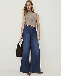 A woman in a taupe cable-knit mock neck top and dark wash wide-leg jeans stands against a plain, light-colored wall, holding a brown suede bag. carousel Product Image 3