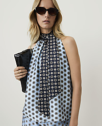 A woman in a light blue patterned halter top and sunglasses holds a black clutch in front of a plain background.