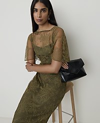 A woman in a green lace dress and holding a black clutch sits on a stool against a plain white background. carousel Product Image 4