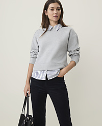 A woman in a grey sweatshirt layered over a striped collared shirt and dark pants stands against a plain, light background. carousel Product Image 1