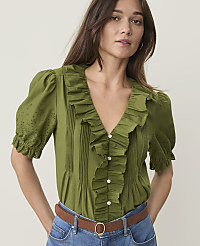 A woman in a ruffled green blouse and blue jeans stands against a plain, light background. carousel Product Image 1