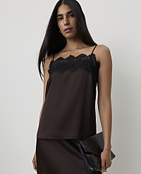 A woman in a brown camisole with black lace trim holds a black clutch in front of a plain background.