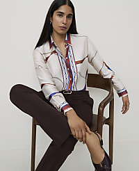 A woman in a patterned silk shirt and dark trousers sits on a wooden chair against a plain background. carousel Product Image 1