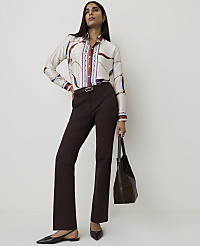 A woman in a patterned blouse and brown pants stands against a plain white background, holding a dark brown bag. carousel Product Image 3