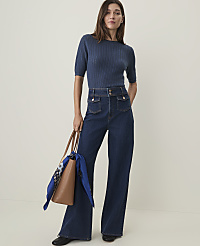 A woman in a blue ribbed sweater and dark wash wide-leg jeans stands holding a brown tote bag with a blue scarf, against a plain, light background. carousel Product Image 1