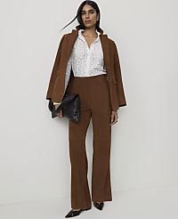 A woman in a brown corduroy suit and a patterned blouse stands against a plain white background, holding a black woven clutch.