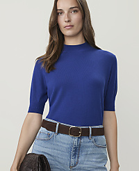 A woman in a blue mock-neck sweater and jeans, accessorized with a brown belt and clutch, stands against a plain beige background. carousel Product Image 1