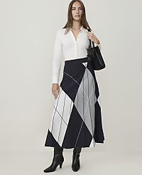 A woman in a white shirt, patterned skirt, and black boots stands against a plain white background, holding a black handbag. carousel Product Image 2