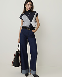 A woman in a sleeveless argyle sweater and wide-leg jeans stands against a plain white background, holding a tote bag. carousel Product Image 1