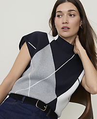 A woman in a sleeveless argyle sweater and dark jeans sits in front of a blurred background.