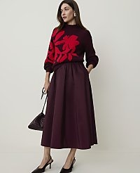 A woman in a burgundy sweater with a red floral print and a matching midi skirt stands against a plain, light-colored background, holding a dark handbag. carousel Product Image 1