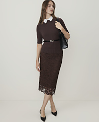 A woman in a brown sweater, white collar, and lace skirt stands against a plain white background, holding a black handbag. carousel Product Image 3