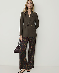 A woman in a brown blazer and patterned pants stands against a plain white wall, holding a small patterned bag. carousel Product Image 3
