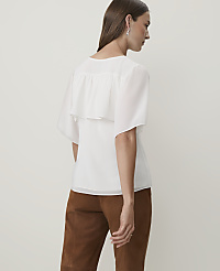 A woman in a white ruffled blouse and brown pants stands against a plain, light background. carousel Product Image 2