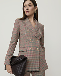A woman in a patterned blazer and pants holds a woven clutch in front of a plain background.