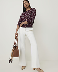 A woman in a patterned burgundy cardigan and white flared pants stands holding a brown handbag against a plain white background. carousel Product Image 3