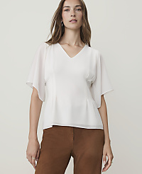 A woman in a white v-neck blouse and brown pants stands against a plain, light gray background. carousel Product Image 1