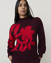 A woman in a maroon sweater with a large red floral design stands against a plain, light-colored background. carousel Product Image 1