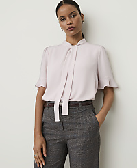 A woman in a light pink bow-neck blouse and plaid trousers stands against a plain white background.