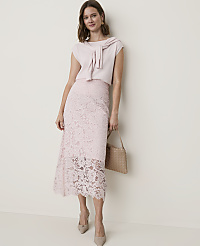 A woman in a light pink lace skirt and matching top stands against a plain, light-colored wall, holding a woven handbag. carousel Product Image 4