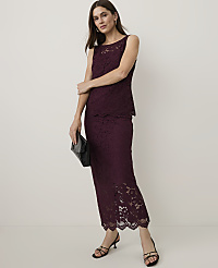 A woman in a burgundy lace dress and black heels stands with a clutch in front of a plain background.