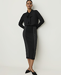 A woman in a black bow-neck blouse and a black pencil skirt with white geometric detailing stands against a plain white background. carousel Product Image 3
