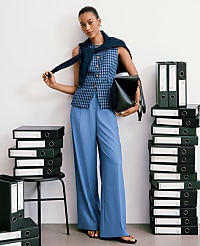 A woman in blue pants and a patterned vest stands between stacks of black binders against a white wall. carousel Product Image 4