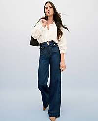 A woman in a white blouse and wide-leg jeans stands against a light blue background, with a dark jacket draped over her shoulder. carousel Product Image 4