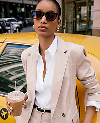 A woman in a tan plaid suit and sunglasses holds a coffee cup while leaning against a yellow taxi in a city street. carousel Product Image 3
